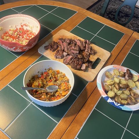 A table laid with sliced beef steak on a board, roast potatoes, sautéed peppers and a tomato salad